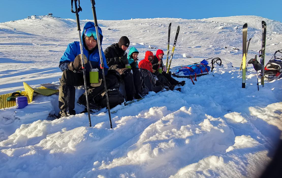 Group sitting in the snow on mountain slopes, enjoying hot drinks during an Arctic ski expedition.