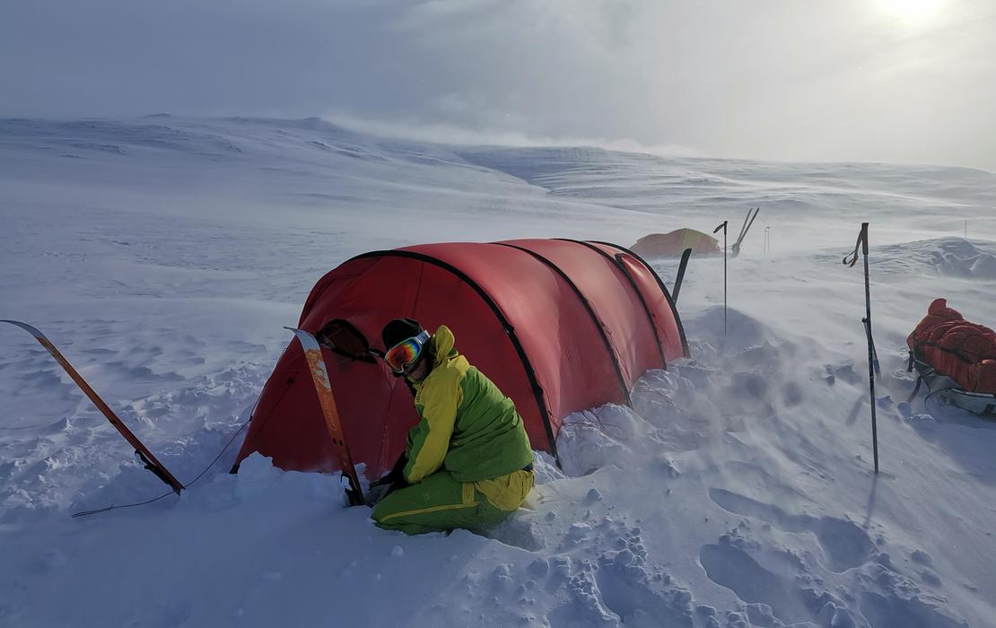 Person crouching beside a tent in a snowy, windy Arctic landscape with skis standing upright nearby.