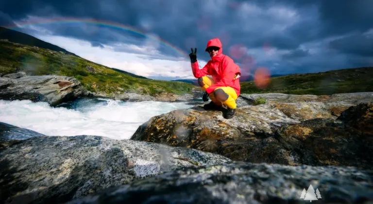 Hiker in Lapland mountains by a stream, rainbow arching over the Arctic landscape.
